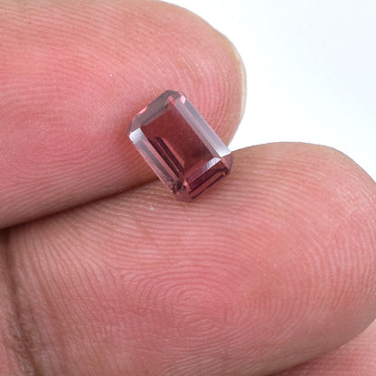 Emerald-cut gemstone with a pinkish-red hue held between fingertips.