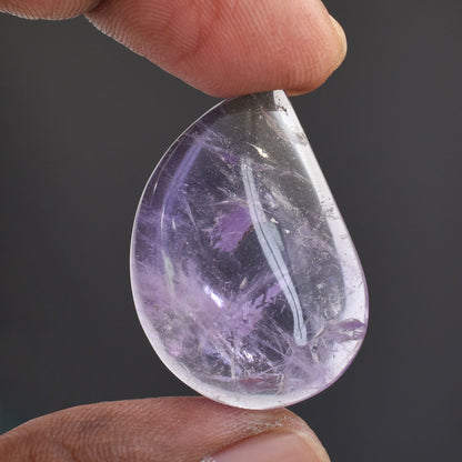 Teardrop-shaped purple crystal or gemstone held between two fingers.