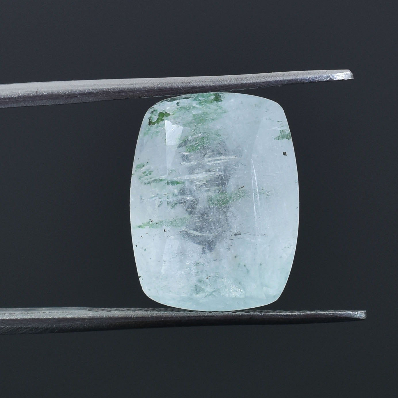 Pale green gemstone with a cushion cut held by tweezers.