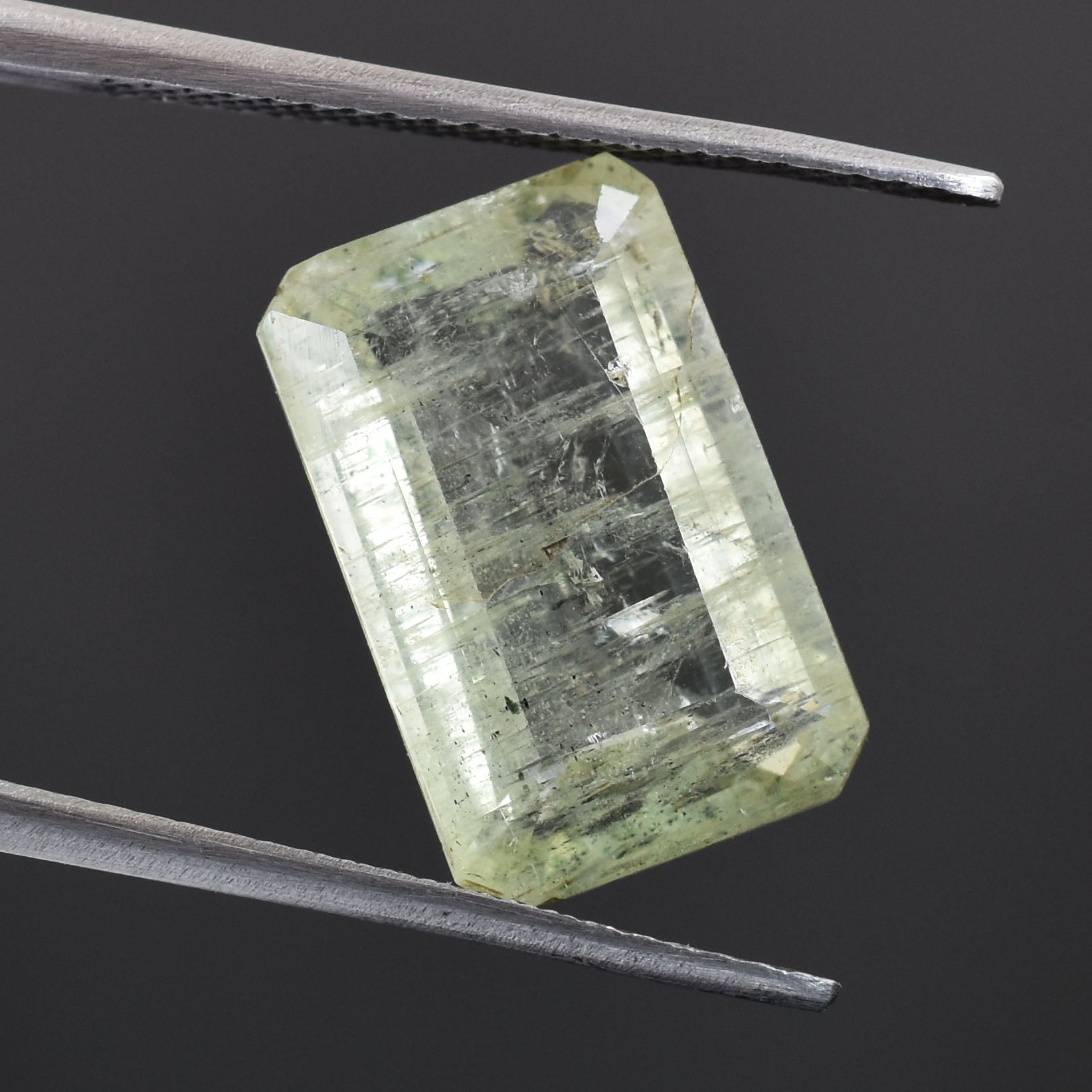 Pale green, rectangular-cut gemstone held by tweezers.