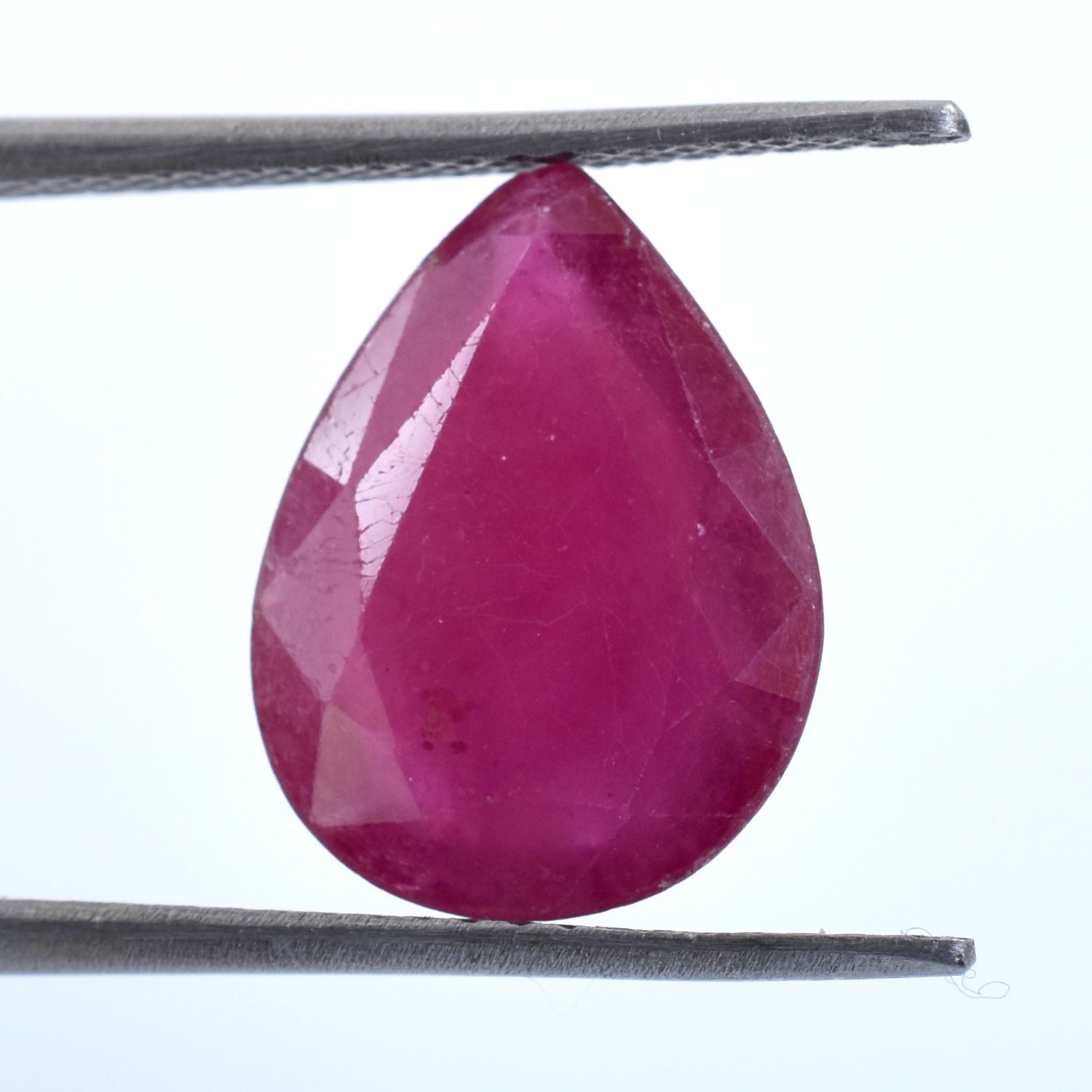 Teardrop-shaped, faceted ruby gemstone held by tweezers.